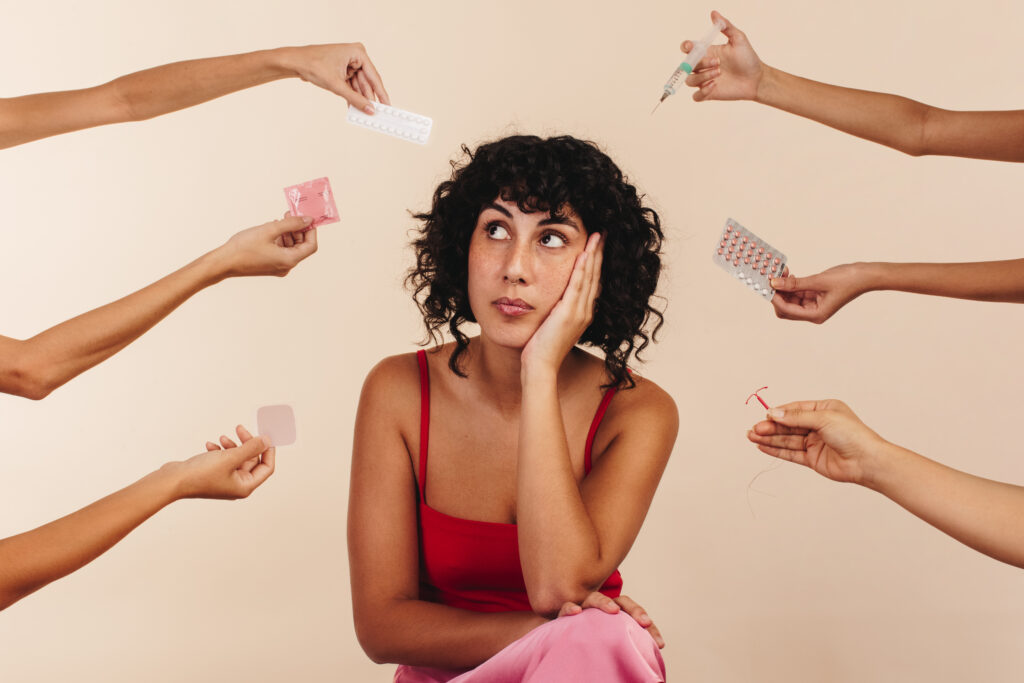 Young woman looking away thoughtfully while surrounded by hands holding different forms of hormonal and non-hormonal contraception. Pensive young woman making a decision about her reproductive health.